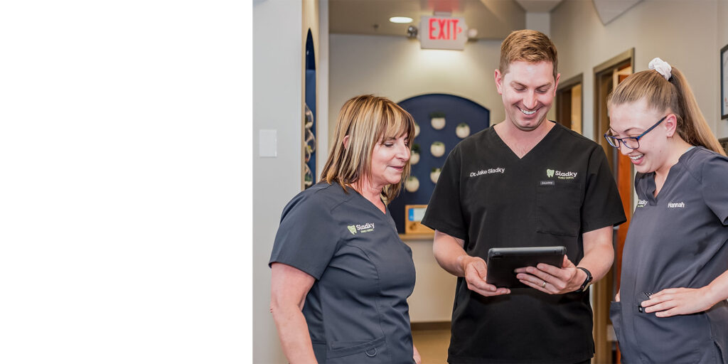 Dr. Jake Sladky, labeled “Chiropractor,” holds a tablet while discussing with two staff members in “Sladky”-branded uniforms, including one named Hannah, in a clinic hallway with circular wall decor.