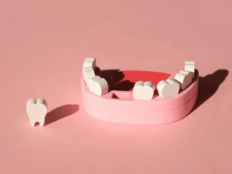 A pink dental model representing the upper jaw with several white teeth. One tooth is placed outside the model on the left side. The background is a solid pink color.